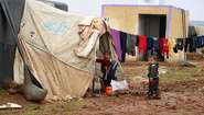 2020-winterhilfe-syrien-kinder-camp-welthungerhilfe.JPG Zwei Kinder stehen vor einem Zelt in einem Flüchtlingscamp und schauen Richtung Kamera. An einer Leine ist Wäsche aufgehängt.