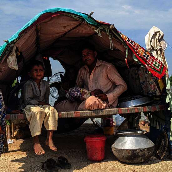 Fluten in Pakistan: Ein Mann, eine Frau, zwei kleine Kinder und ein Baby in ihrer Notunterkunft, die aus Holzstäben, Decken und einer Plastikabdeckung besteht. im Hintergrund ist Wasser zu sehen.