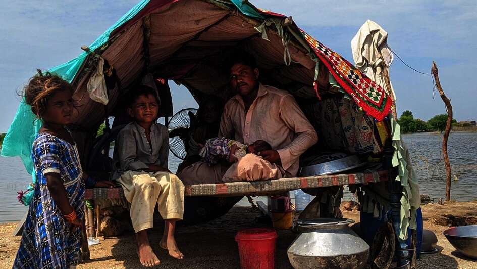 Fluten in Pakistan: Ein Mann, eine Frau, zwei kleine Kinder und ein Baby in ihrer Notunterkunft, die aus Holzstäben, Decken und einer Plastikabdeckung besteht. im Hintergrund ist Wasser zu sehen.