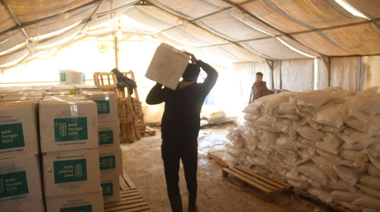 2019-nahrungsmittelhilfe-nord-aleppo-verteilung-syrien_C_welthungerhilfe.jpg Im Innern eines Zeltes: Links stehen aufgereihte und gestapelte Pakete der Welthungerhilfe. In der Mitte ein Mann von hinten, der ein Paket auf seinen Schultern trägt.