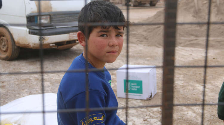 2019-pressefoto-geberkonferenz-syrien-fluechtlingscamp-azzaz-junge_C_welthungerhilfe.JPG Ein Junge sitzt hinter einem Gitter und schaut in die Kamera. Im Hintergrund sieht man ein Welthungerhilfe-Paket.