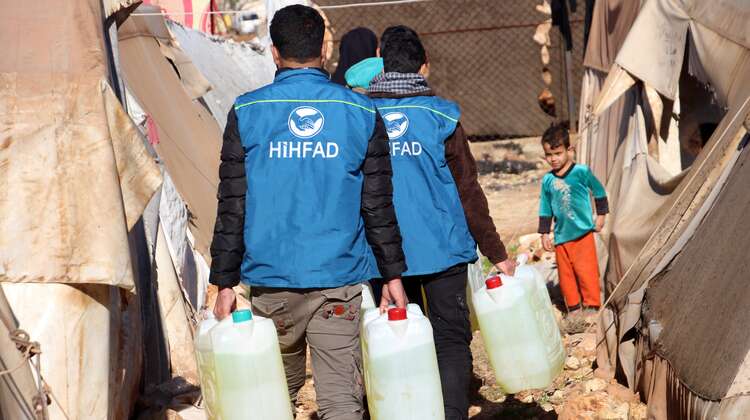 2020-winterhilfe-syrien-diesel-verteilung-hihfad-kind-welthungerhilfe.JPG HIHFAD-Mitarbeiter in blauen Westen tragen Diesel in Kunststoffkanistern zu einem Zelt. Ein Kind schaut ihnen entgegen.