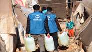 2020-winterhilfe-syrien-diesel-verteilung-hihfad-kind-welthungerhilfe.JPG HIHFAD-Mitarbeiter in blauen Westen tragen Diesel in Kunststoffkanistern zu einem Zelt. Ein Kind schaut ihnen entgegen.
